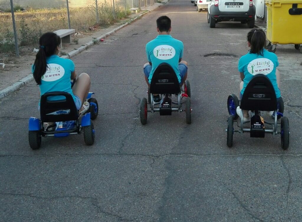 Tres monitores de i-animese, de espaldas y con camiseta azul, sobre karts a pedales en la calle
